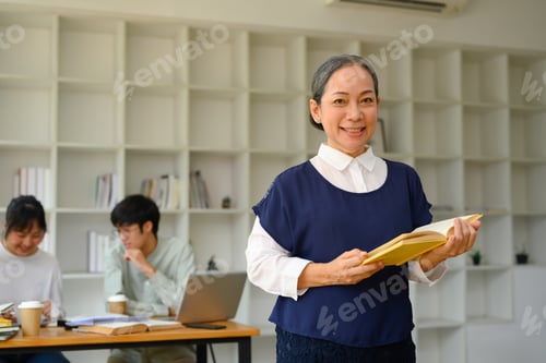 Preview: Portrait of friendly smiling mature professor holding book standing in class