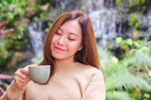 Preview: A young woman holding and drinking coffee while sitting in the garden with waterfall