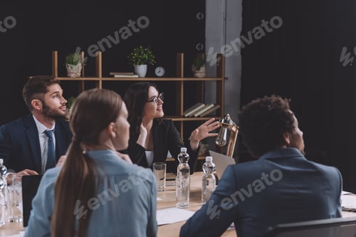 Preview: young businesswoman talking during business meeting while sitting near multicultural colleagues