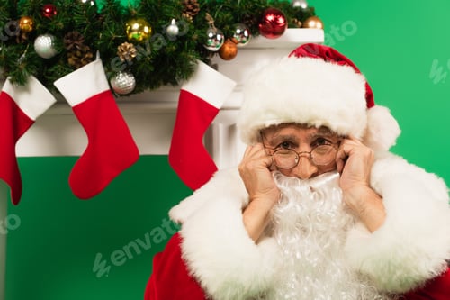 Preview: Upset santa claus looking at camera near decorated fireplace on green background