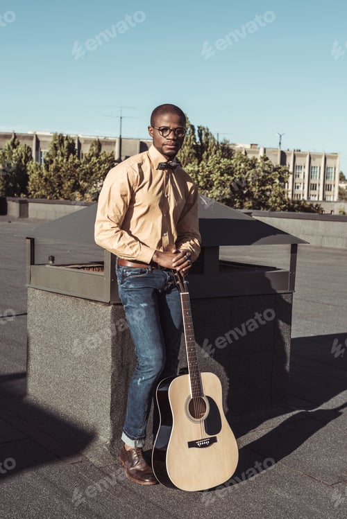 Preview: Young african american man in eyeglasses, posing with acoustic guitar on roof of building
