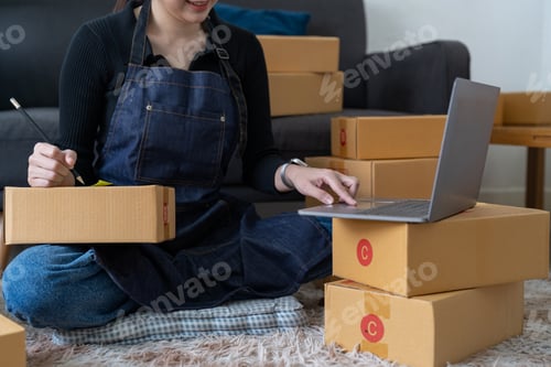Preview: startup small business owner writing address on cardboard box at workplace. Freelance Asian woman