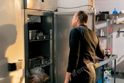 Preview: chef stands in kitchen in a black uniform and looks through what is on the list in the refrigerator