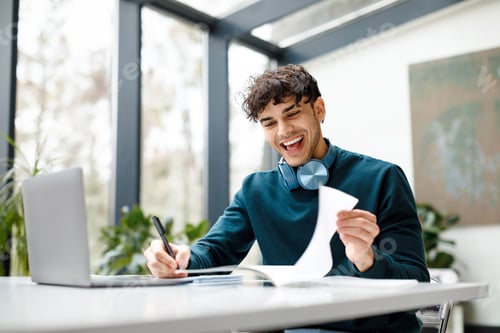 Preview: Happy student guy using laptop computer learning online, taking notes and checking copybook during