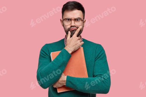 Preview: Displeased unshaven young man holds chin, purses lips, has serious unhappy facial expression, holds