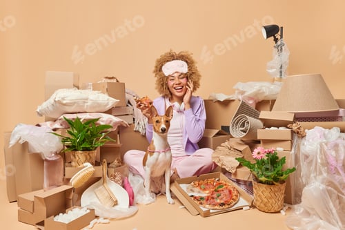 Preview: Positive curly haired woman moves to new house eats delicious pizza poses near pedigree dog celebrat