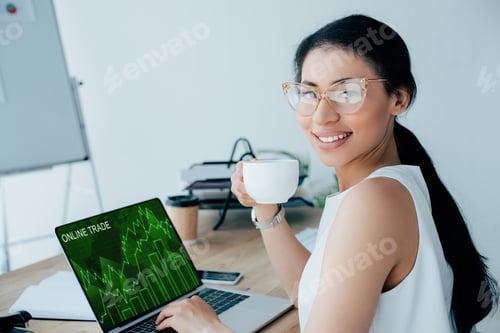Preview: smiling latin businesswoman using laptop with online trade website on screen while holding coffee