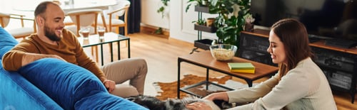 Preview: Smiling woman petting border collie dog on couch near boyfriend in living room, banner
