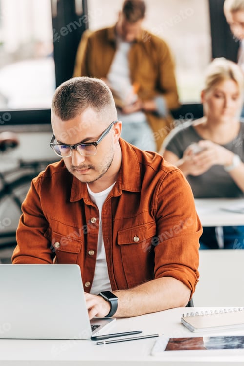 Preview: handsome young man working with laptop at open space office with colleagues on background