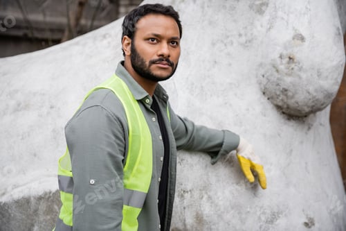 Preview: Serious indian worker in high visibility vest and glove standing near concrete sculpture in outdoor