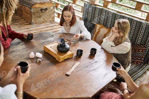 Preview: woman reading tarot cards near multiethnic girlfriends and tea on wooden table in cozy house