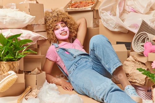 Preview: Happy tired curly haired woman applies beauty mask on face wears t shirt and denim overalls lies aro