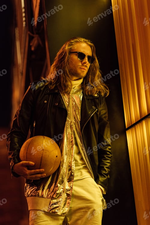 Preview: stylish young man in sunglasses and leather jacket holding golden basketball ball under yellow light