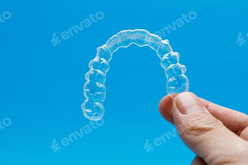 Preview: Hand holding one transparent retainer on blue background. Invisalign orthodontics concept