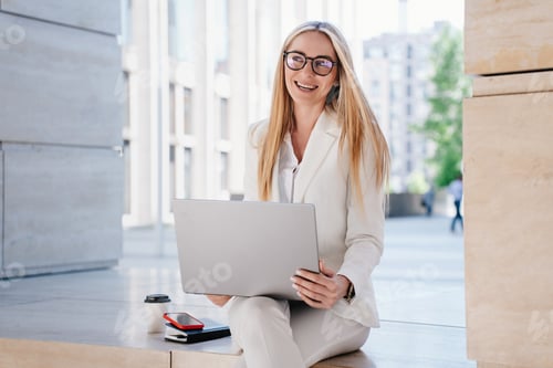 Preview: young woman in glasses and white suit outdoors working with laptop, looking aside with broad smile