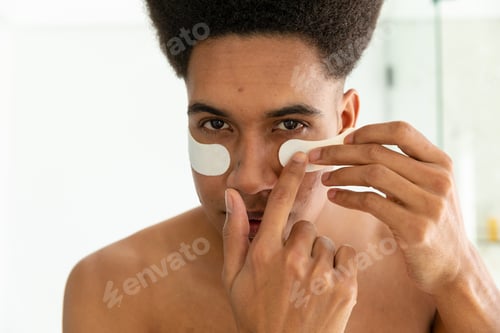 Preview: Biracial man looking in mirror and applying under eye masks in sunny bathroom