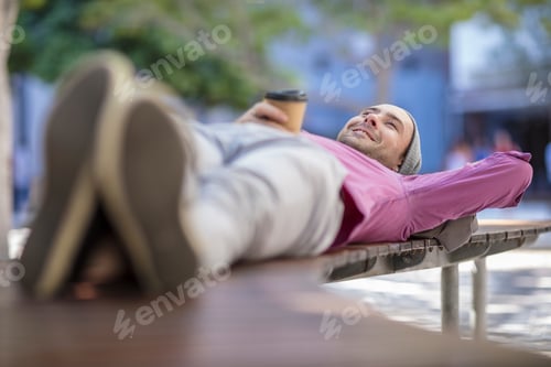 Preview: Young man outdoors, lying on bench, holding takeaway coffee cup