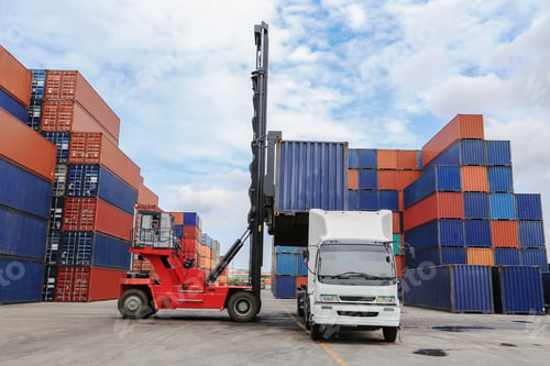 Vista previa: A red container handler loads a shipping container onto a white truck in a busy container yar