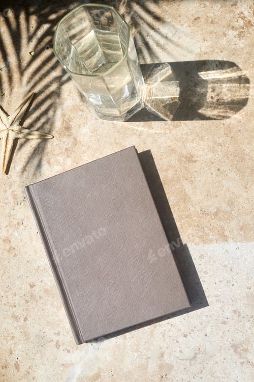 Preview: Summer Reading Setup. A brown book and a glass of water on a sunlit table with palm shadows.