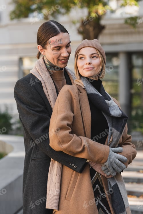 Preview: tattooed man hugging pleased blonde woman in hat while standing outside