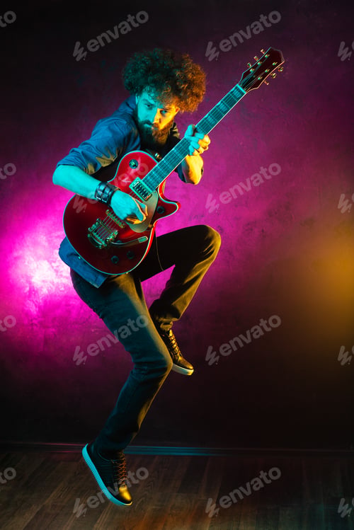 Preview: Bearded hipster man with curly hair jump with red guitar in neon lights.
