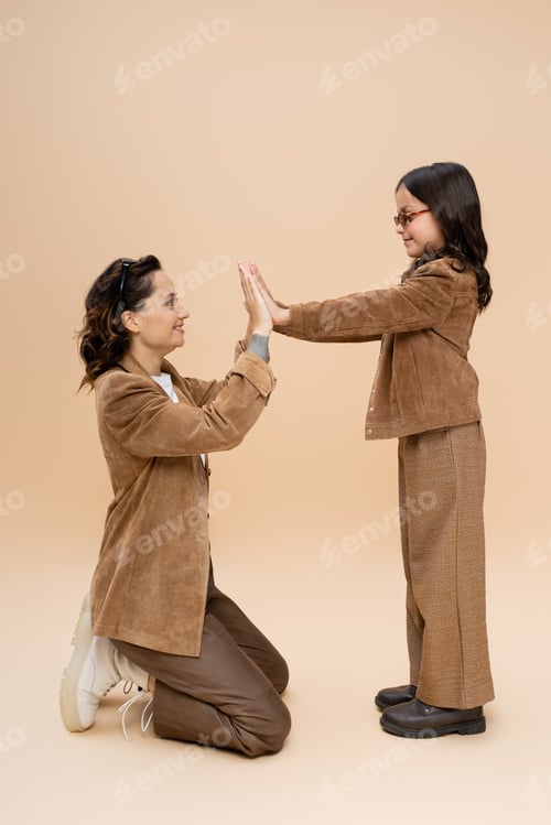 Preview: side view of mother and daughter in stylish autumn outfit playing patty cake game on beige