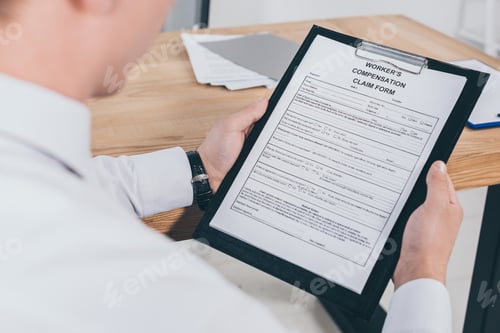 Preview: cropped view of adult businessman reading compensation claim form at workplace