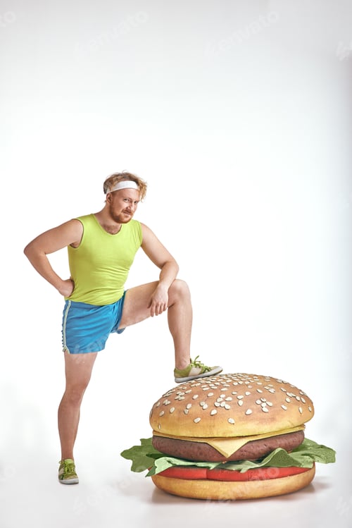 Preview: Red haired, bearded, plump man put his leg to a huge sandwich