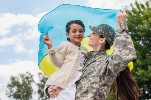 Preview: cheerful military woman with daughter holding ukrainian flag outdoors