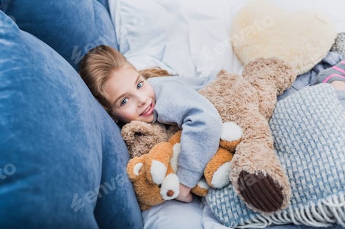 Preview: Adorable little girl lying on bed with teddy bears and smiling at camera