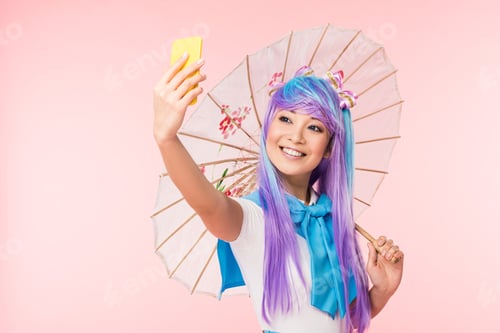 Preview: Smiling asian anime girl with paper umbrella taking selfie isolated on pink