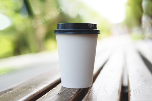 Preview: A paper cup of coffee to go on bench with blurred nature and sun background.