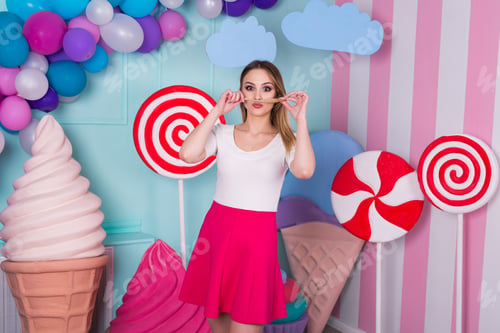 Preview: Portrait of joyful young woman in pink dress on background decorated with huge candies and ice cream