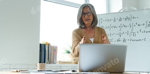 Preview: Mature woman teaching mathematics while standing near the whiteboard and looking at laptop