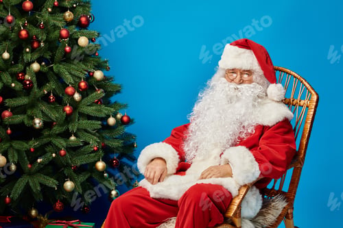 Preview: cheerful Santa Claus with beard and eyeglasses sitting in rocking chair near Christmas tree on blue