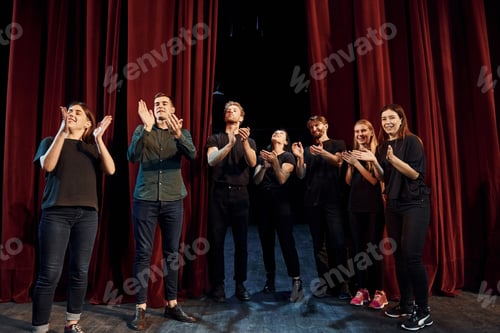 Preview: Standing against red curtains. Group of actors in dark colored clothes on rehearsal in the theater