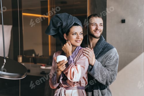 Preview: Cheerful young woman in silk gown and towel wrapped around head holds jar of skin care cream