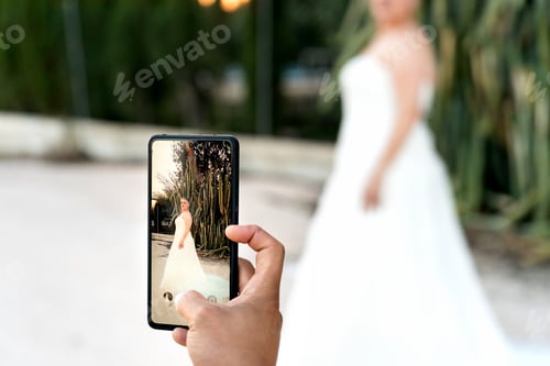 Preview: Photographer capturing bride's elegance on smartphone during wedding photo shoot