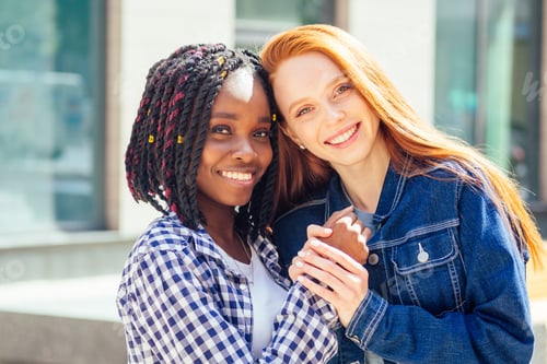 Preview: cheerful afro and european lesbian women laughing and looking at camera at summer downtown city