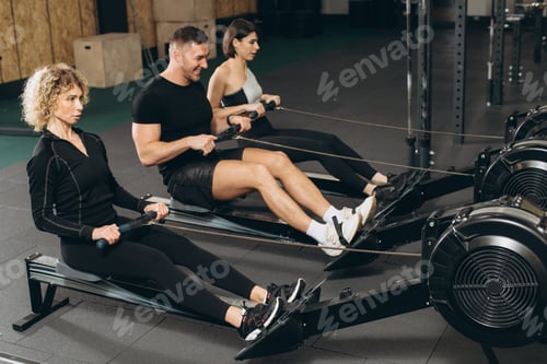 Preview: Young man and beautiful women working out with rowing machine at crossfit gym.