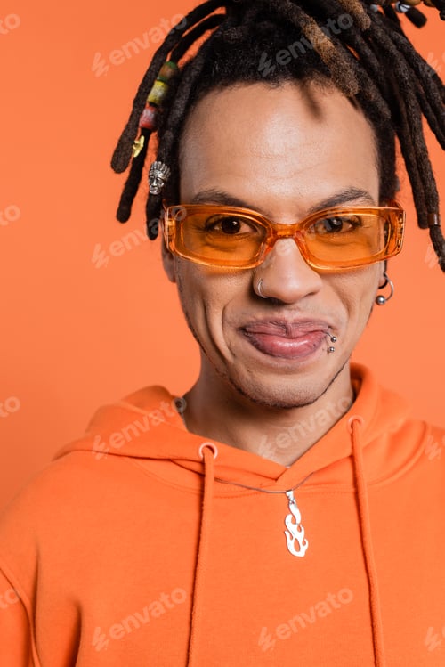 Preview: portrait of satisfied multiracial man with dreadlocks wearing stylish sunglasses isolated on coral