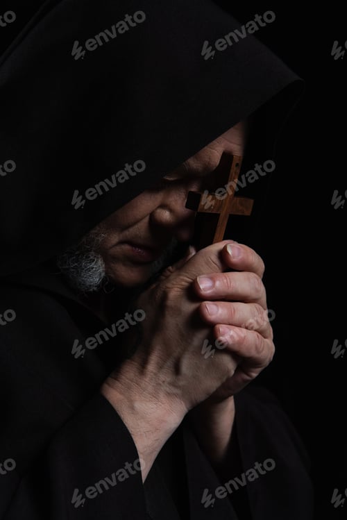 Preview: mysterious priest in hood obscuring face with crucifix while praying isolated on black