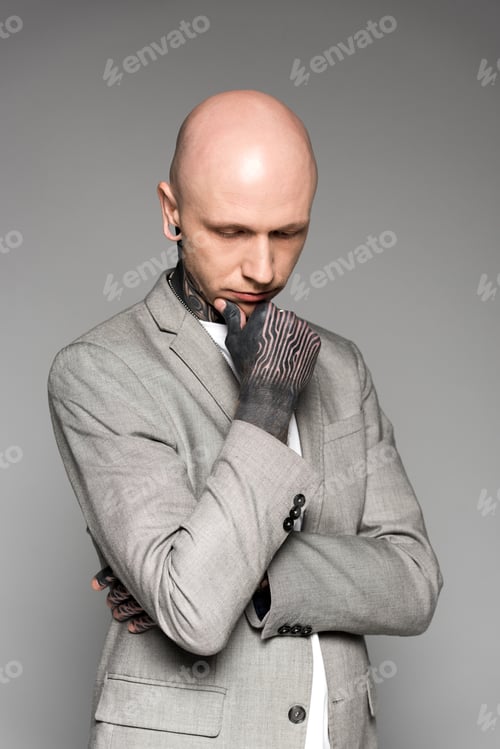 Preview: young tattooed man in suit standing with hand on chin and looking down isolated on grey