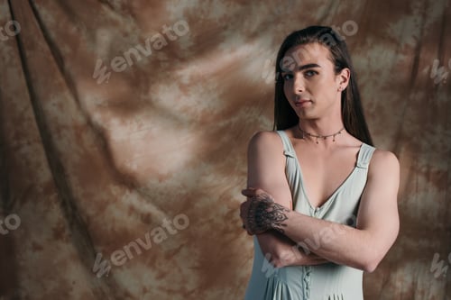 Preview: Young queer person in sundress looking at camera on abstract brown background