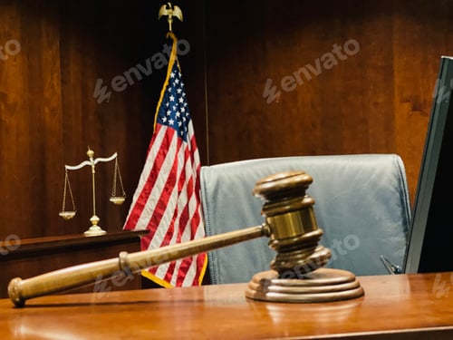 Preview: Judge’s wooden gavel on judicial bench, scales of justice and flag of US in background.