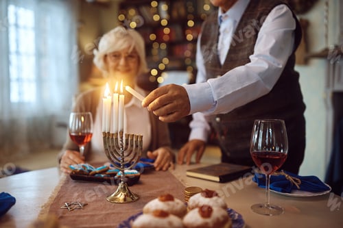 Preview: Close up of mature man lighting menorah candles while celebrating Hanukkah with his wife at home.