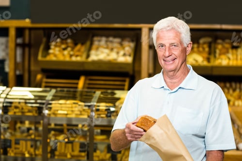 Preview: Senior man smiling and holding loaf of bread in paper bag inside bakery shop, copy space