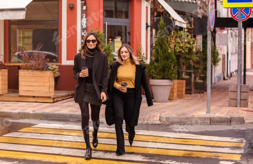 Preview: Two elegant female friends in trendy clothes drinking coffee and talking while walking together