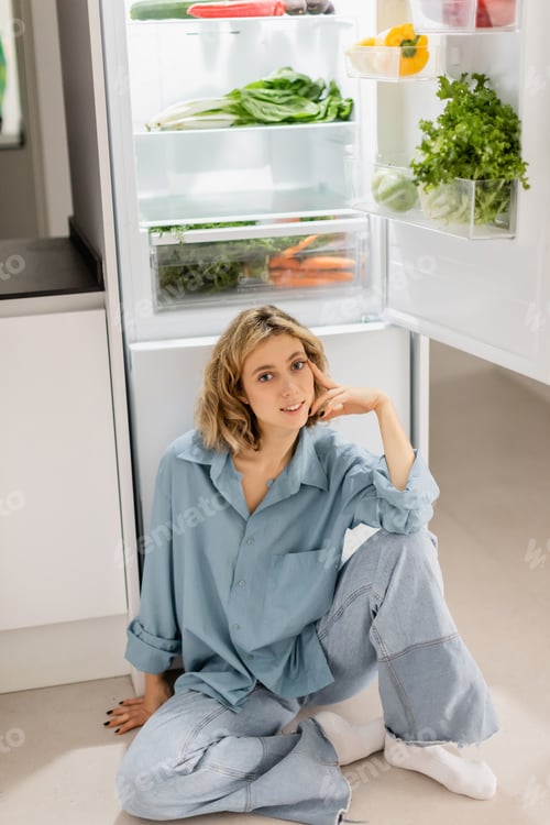 Preview: happy young woman sitting near opened refrigerator with fresh products in kitchen