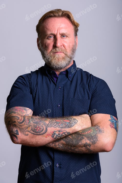 Preview: Mature bearded man with hand tattoos with arms crossed in gray b